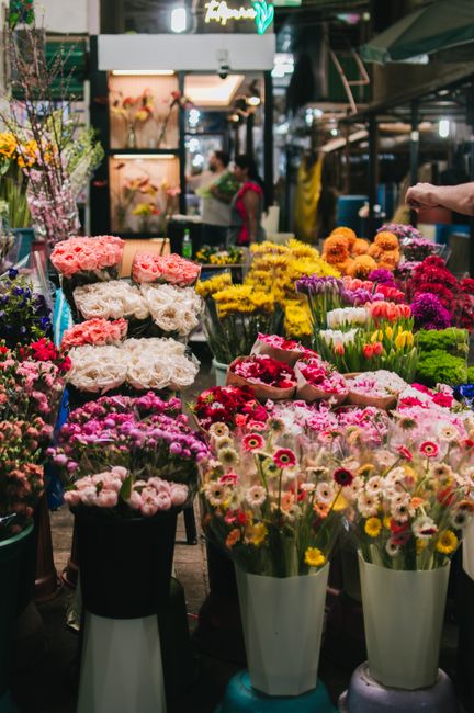 A bustling flower market scene featuring a diverse array of fresh flowers arranged in various vases and bouquets. Prominent in the foreground are cheerful daisies, tulips, and chrysanthemums in shades