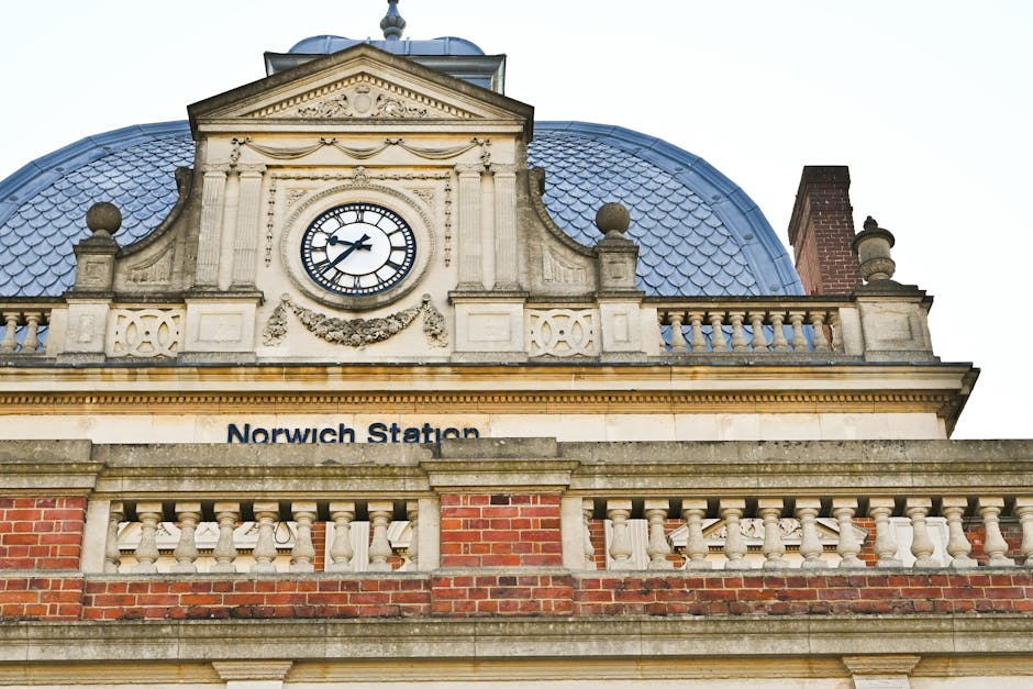 A detailed view of Norwich Station's historic facade featuring a central clock with black Roman numerals and hands, framed by ornate stonework and decorative garland carvings. The building displays a 