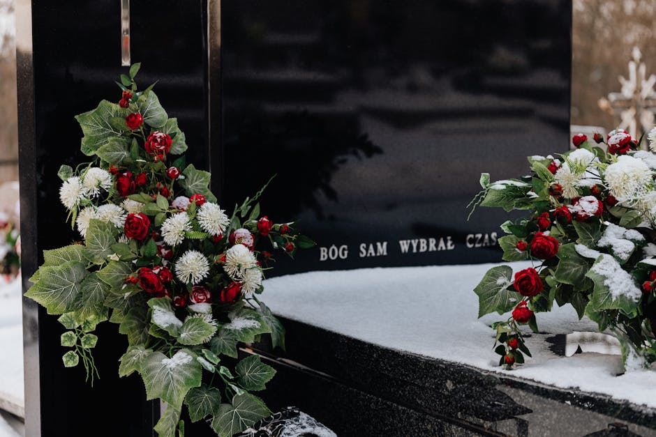 A black granite gravestone adorned with two lush floral arrangements, featuring red roses, white chrysanthemums, and sprigs of green ivy. The flowers are arranged in cascading bouquets, with some flow