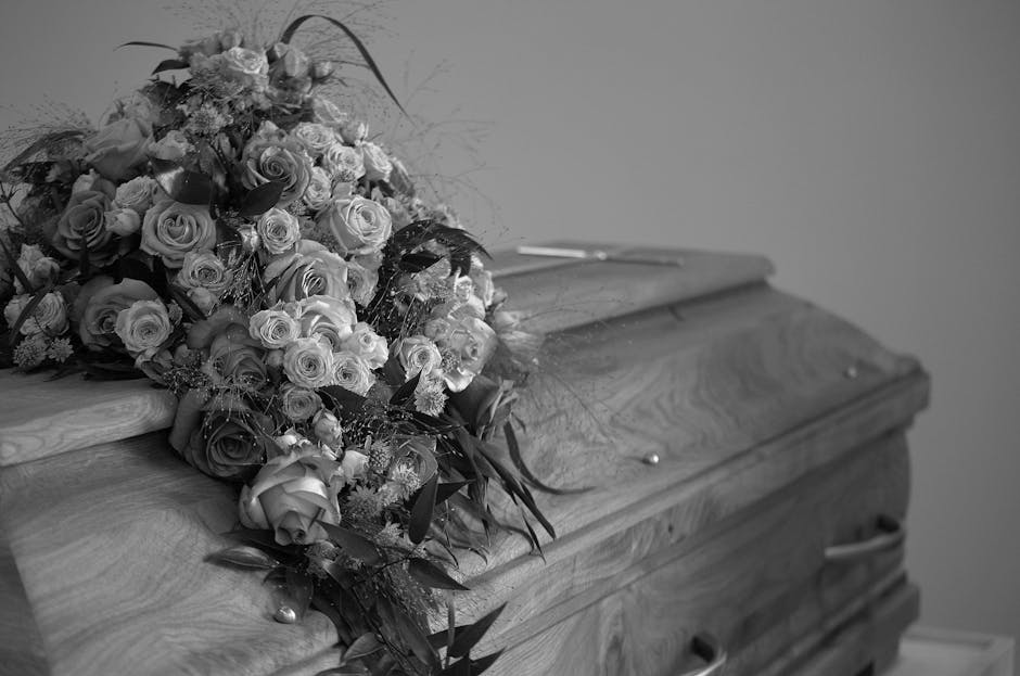 A large, elegant floral arrangement resting on top of a wooden casket, featuring a variety of fresh roses in shades of cream, soft pink, and deep red, complemented by greenery and delicate filler flow