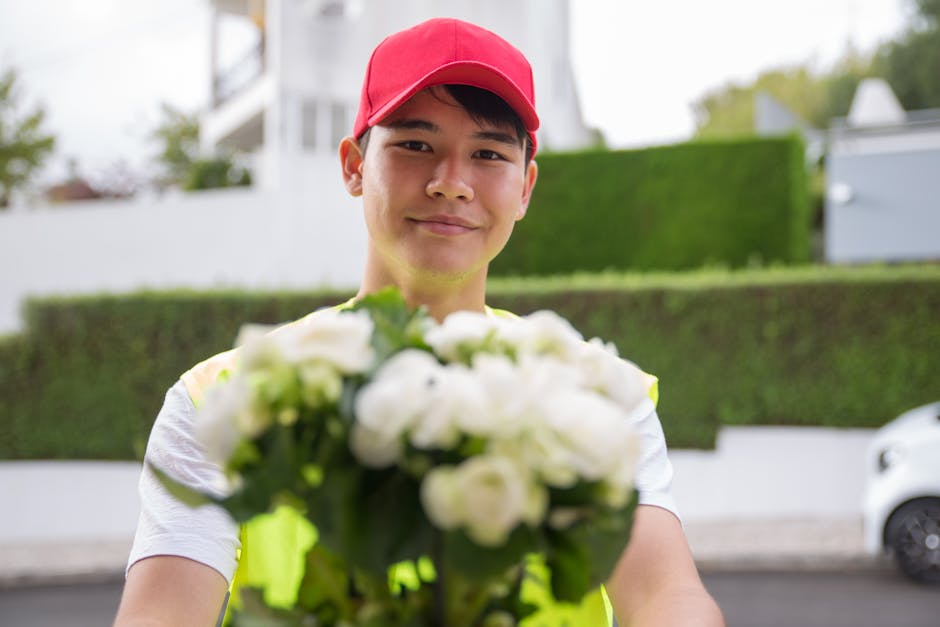 A delivery person wearing a red cap and a high-visibility vest is smiling and holding out a vibrant bouquet of fresh white flowers, possibly roses or peonies, wrapped with green foliage. The bouquet i