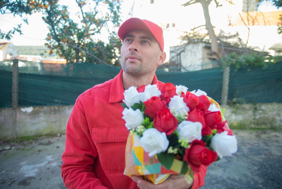 A man wearing a red cap and a matching red jacket is holding a floral bouquet wrapped in colorful paper, featuring red and white roses with green foliage. The bouquet appears fresh with vibrant petals