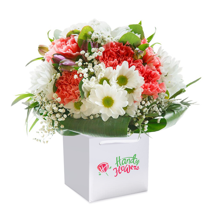 Presented in a modern square white gift bag, this bouquet is a vibrant celebration of freshness from the heart of Enfield Town. Coral and soft peach carnations, each one abundant with characteristic ruffled petals, form the lush center-calling to mind the inviting blooms you might discover near Enfield Town's bustling High Street or the peaceful green of St. Andrew's Church gardens. Interspersed amid the spectacular carnations are snowy white daisies with striking lime-green centers, infusing the arrangement with a cheerful brightness reflective of the community's spirit. Sprays of light and airy baby's breath weave between the larger flower heads, imparting a touch of gentle sophistication as soft as a morning in Bush Hill Park. Peeking out on the left, you'll find a few purple-tinged buds-possibly alstroemeria yet to open-hinting at the promise of more beauty to come. Deep green foliage, finished with broad, glossy leaves, forms an elegant collar that both frames and elevates the bouquet's rounded design. The entire arrangement is set neatly within a structured white bag, complete with white ribbon handles, made for easy gifting or delivery from Enfield Town Flowers throughout local neighbourhoods. The white-on-white photography and natural, gentle lighting further highlight the bouquet's freshness and poise, making it perfect for birthdays, anniversaries, or as a thoughtful gesture for friends and family in Enfield Town. This image reflects the care, artistry, and commitment of a trusted local florist.
